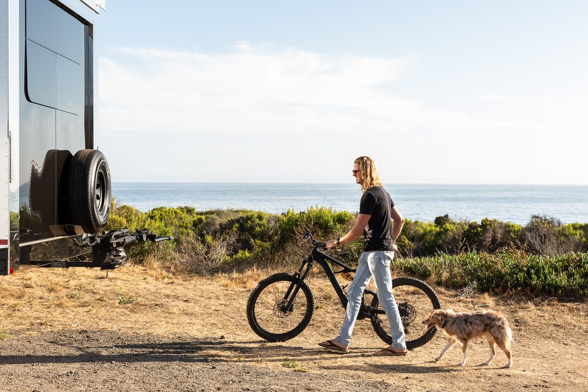 An owner walks a bike and a dog past a Living Vehicle on the coast