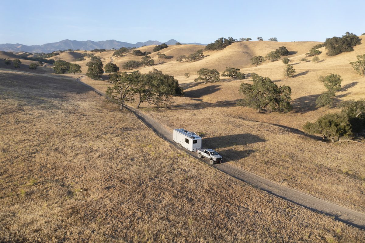 A truck tows a Living Vehicle HD29 along a road through golden hills, seen from above